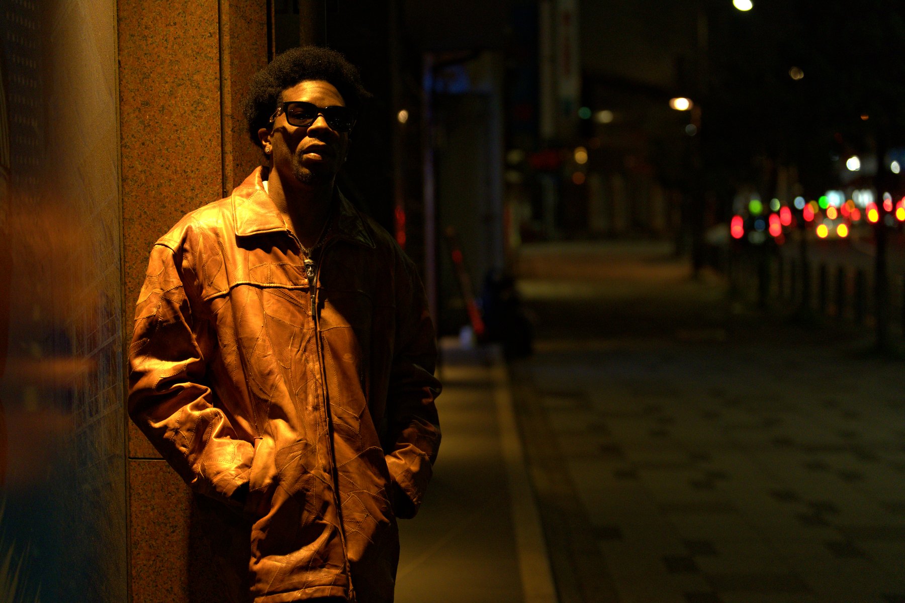 Night portrait of a man in a leather jacket against a city wall, street lights glowing behind — photograph by Wayne Anderson